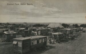 A black and white postcard image of wooden chalets arranged in rows in a field near the sea. There is a large marquee in the background. The title of the postcard reads Central Park, Seton Sands.