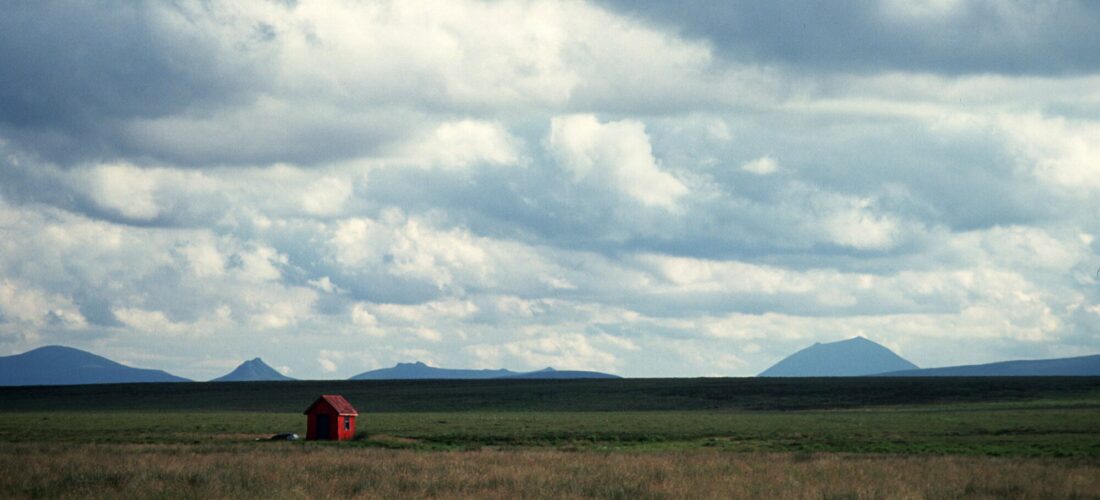 A small red hut with a sloping roof sits alone in a wide open grassy area, with mountains and blue sky in the distance.