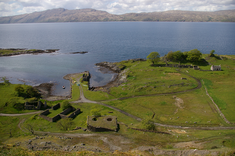 The remains of a harbour, stone buildings and roadways which once formed An Sailean lime works. They are beside the sea on an island. Hills on another island can be seen across the water. 