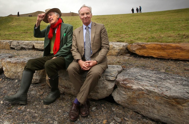 Charles Jencks and the Duke of Buccleuch sitting on a stone at the Crawick Multiverse. Jencks wears a green jacket, green wellies, a red scarf and a brimmed hat. The Duke is in a brown suit and tie. 