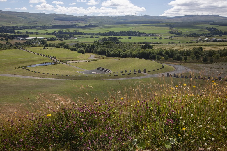 A view across the amphitheater and standing stones at Crawick Multiverse and the rolling green hills beyond. There are wildflowers in the foreground. 