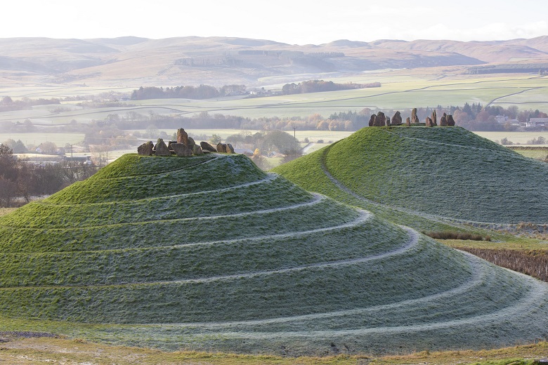 Two grassy mounds topped with collections of standing stones. Narrow paths lead up to the top of the mounds in a circular fashion.
