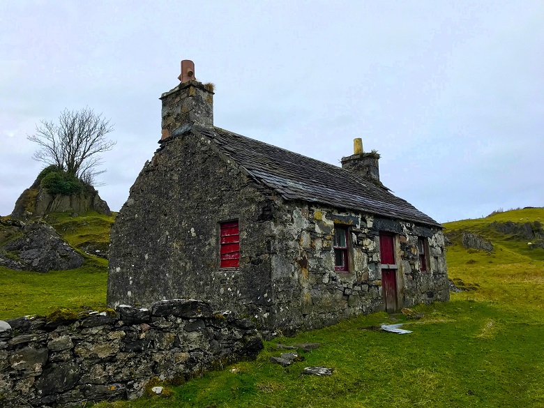 A small stone cottage with a red door, two small windows and a chimney at each end. While it appears to be abandoned, it is not entirely ruined.