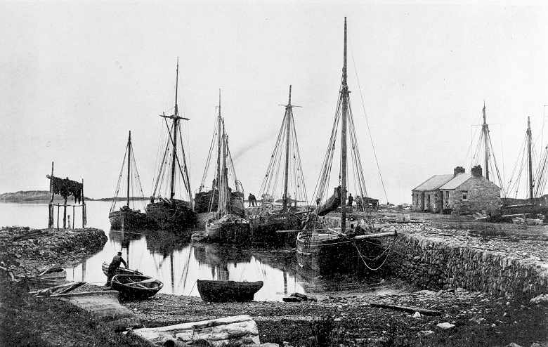 A black and white archive photo of six vessels docked at a small harbour. A figure is working beside a smaller rowing boat in the foreground. 