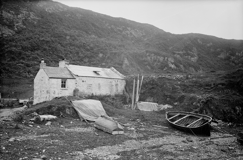 A black and white archive photo of a modest stone house and outbuildings. It is very close to the sea, with a wooden rowing boat and other detritus in front of it. 