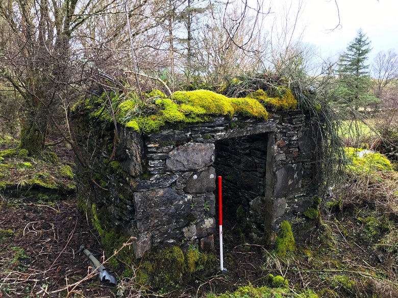 A small and sturdy cube-shaped stone building. It has lost its roof and is covered in bright green moss. 