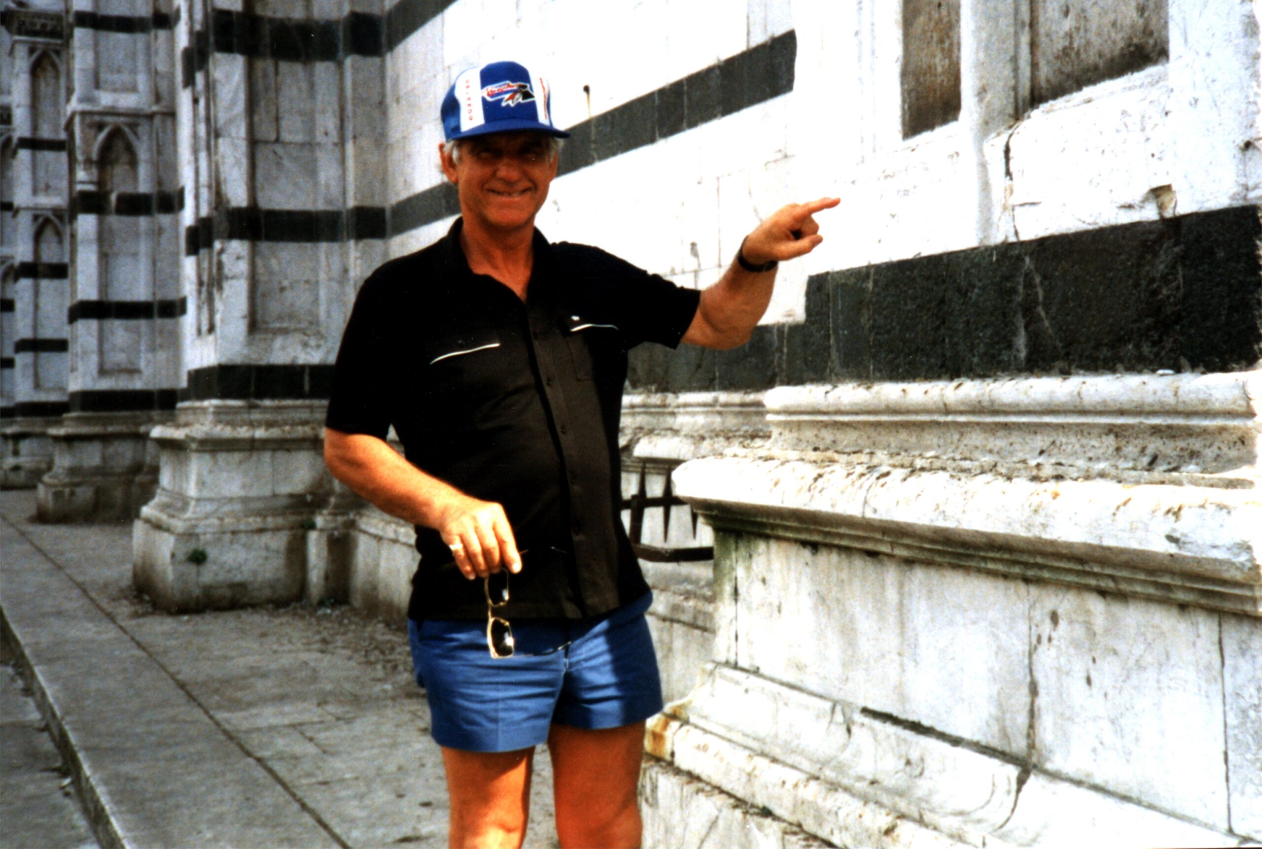 A colour archive photo of Ugolini wearing a polo shirt, shorts and a baseball cap smiling at the camera. It looks like a holiday photo. 