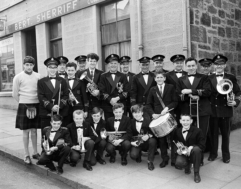 A band made up of men and boys of all ages posing cheerily in their uniforms with their instruments including trumpets, trombones and drums.