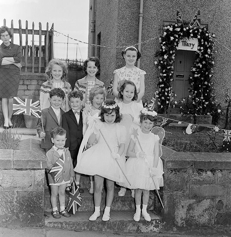 A group of children standing on some garden steps. Two are dressed as fairies, wearing wings and clutching magic wands. Others are very smartly dressed and are holding small union flags. 