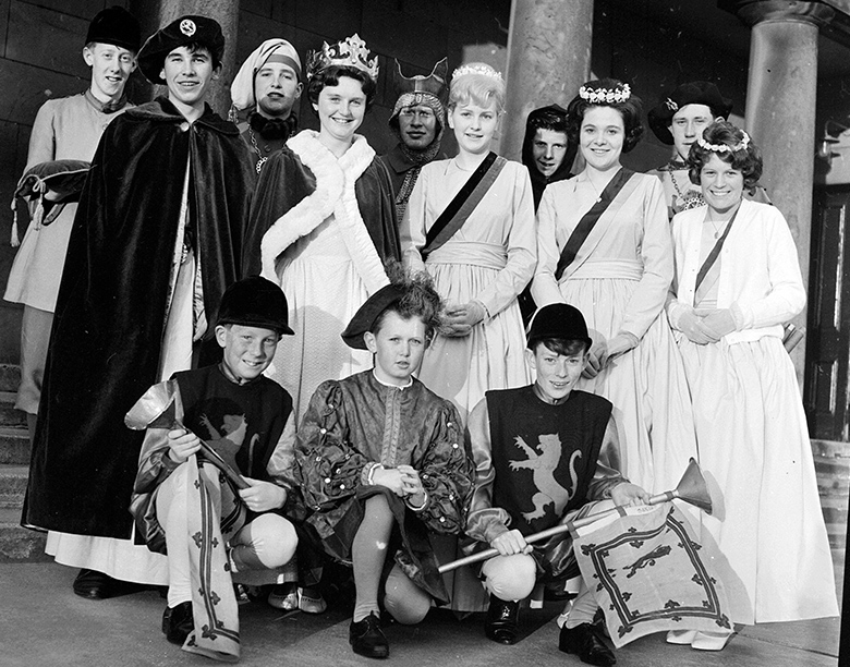 Archive photo of a group of around a dozen young teenagers dressed in medieval and renaissance regalia. The girls wear crowns and sashes, the boys wear cloaks and page boy outfits.