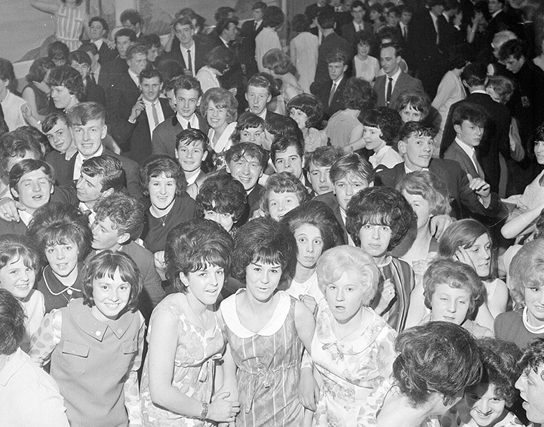 Young people with distinctive 1960s haircuts look up at a camera from a packed dance floor