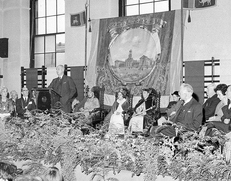 An archive photo of a gala event in a school or community hall. A man is giving a speech from behind a lectern. Others dressed in regal costumes are sitting on wooden thrones. 
