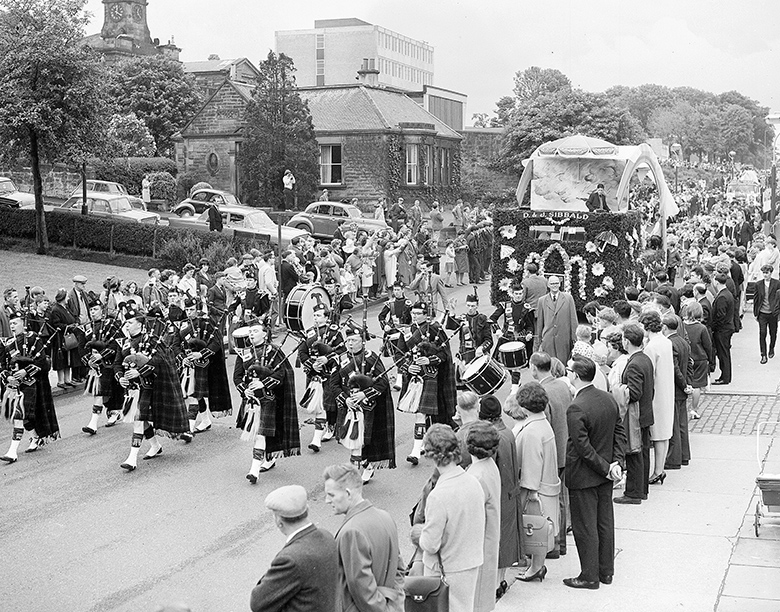 An archive photo of spectators lining both sides of a town street as a pipe band marches past, followed by a lorry which has been lavishly decorated with flowers to create a carnival float.
