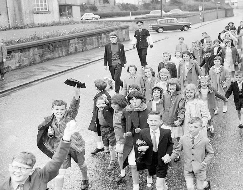 An archive photo of a procession of well-dressed primary school aged children supervised by a teacher and a policeman. 