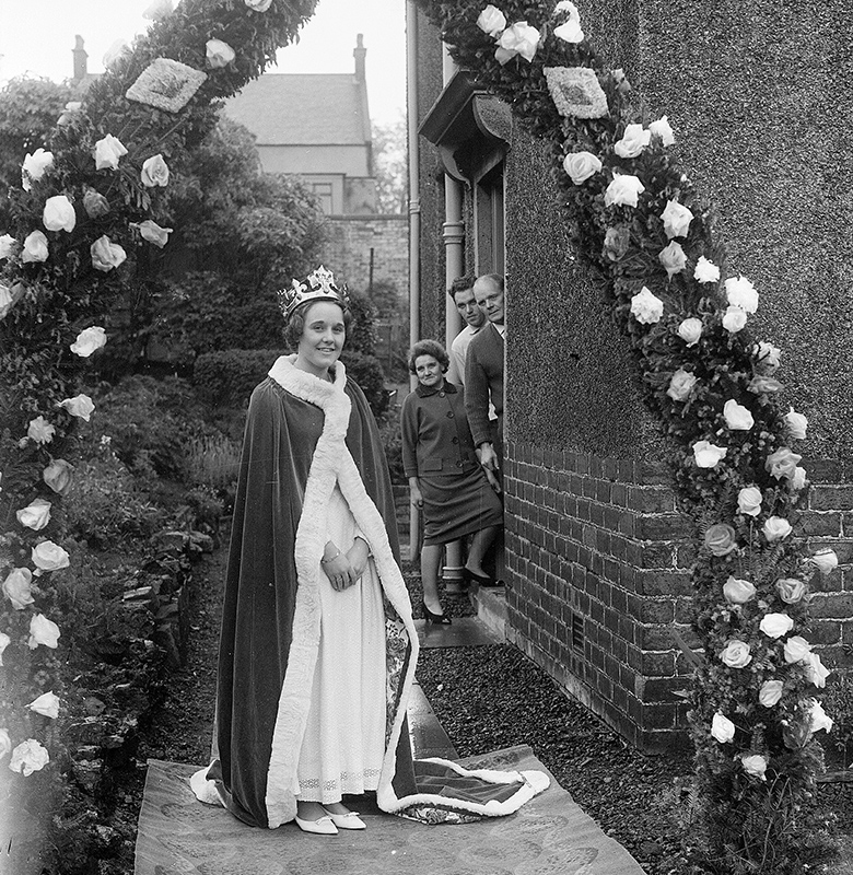 A young lady dressed in a regal-looking velvet robes and a golden crown leaves her house via a arch decorated with flowers. Her family are leaning out of the door.