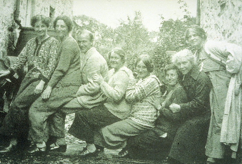 A group of six women and one man pose for a photo. They're having fun and everyone sits on someone else's lap in a big chain.