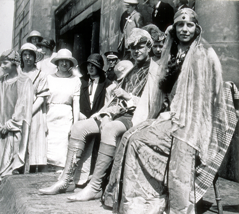 A young woman and a young man wear medieval fancy dress. They sit on a small stage and women in 1920s clothing look on.