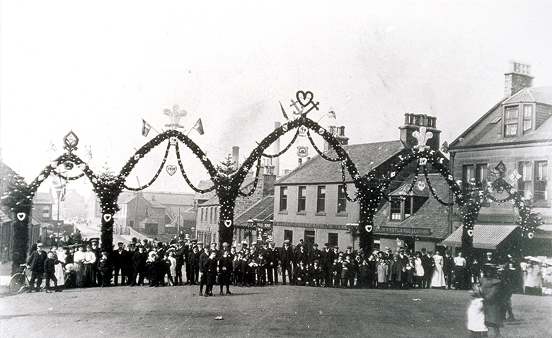 A historic photograph showing townspeople posing under five enormous temporary arches decorated with flags and flowers. They have been erected across a town street.