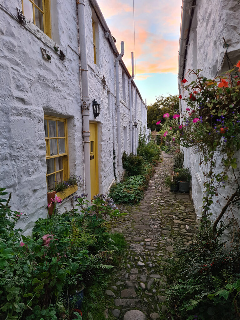 Two rows of small terraced cottages face each other on either side of a narrow cobbled path. The door and window closest to us are painted bright yellow and flowers are planted along the verges.
