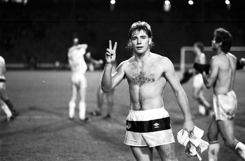 A black and white archive photo of footballer Ally McCoist after a football match. He has taken his shirt off and is holding up two fingers. Other players are swapping shirts in the background.