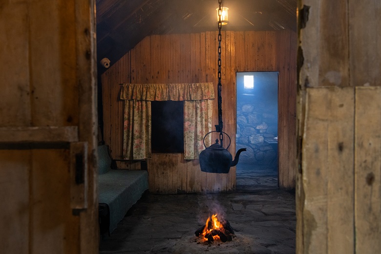 Inside a preserved historic house where a crofting family once lived. There is an open fire in the middle of the room with a black kettle suspended above it. 