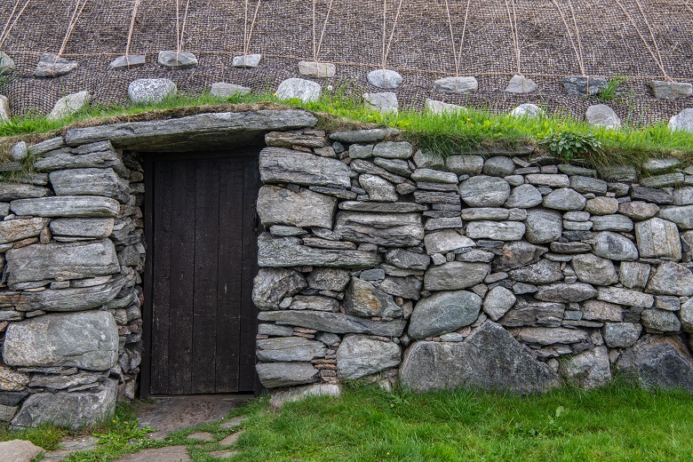 A wooden doorway leading into a small house with stone walls and a thatched roof