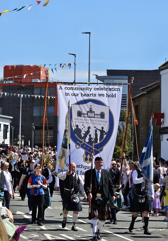 A modern day Gala Day parade being led by flag bearers. A large banner reads "a community celebration our hearts we hold"