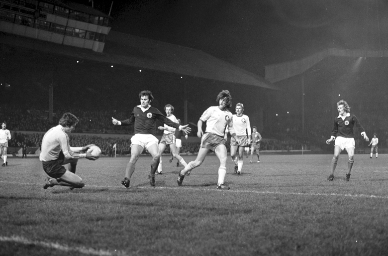 A black and white archive photo of action from a football match. The Scottish team is playing in blue and the Swiss team wears white. Floodlights illuminate the players and the pitch, but the stands and crowd are in the dark.