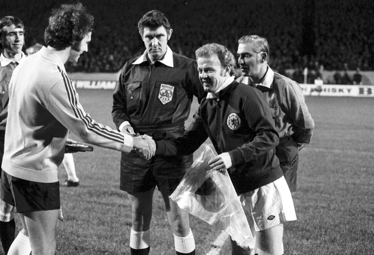 A black and archive photo of two team captains shaking hands and exchanging a pennant before a football match. The referee looks on, wearing a typical 1970s kit with a massive collar.