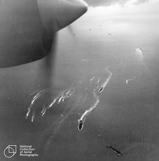 A black and white aerial photo of various landing craft making their way across the sea. Part of the propeller of the plane from which the photo was taken is in the top corner of the image.