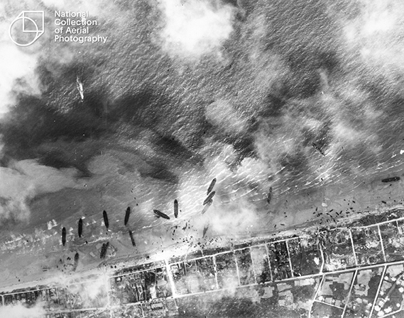 An archive aerial image of landing craft reaching the shoreline of a beach, in quite a haphazard manner. There is smoke above the beaches, presumably the result of heavy gun fire. 