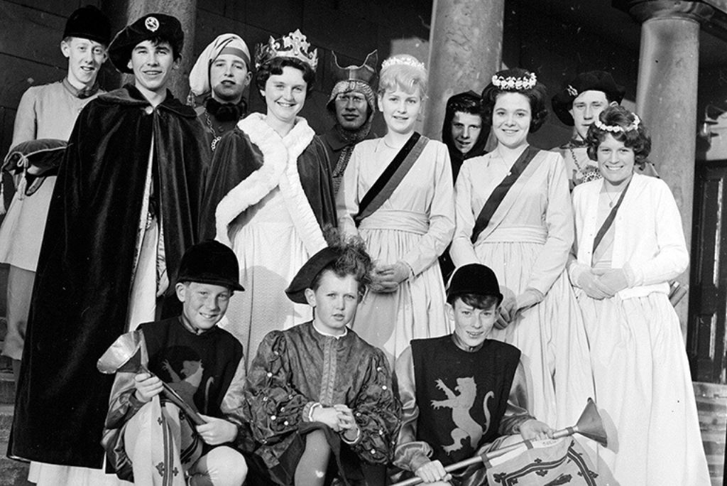 Archive photo of a group of around a dozen young teenagers dressed in medieval and renaissance regalia. The girls wear crowns and sashes, the boys wear cloaks and page boy outfits.