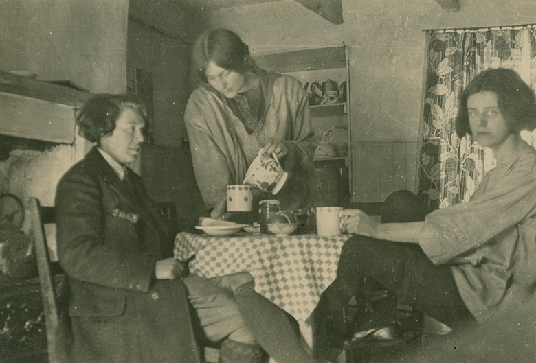 Three women around a table in a small, homely cottage. Dorothy Johnstone is standing and pours tea from a tea pot. The woman at the front left of the image is Vera Jack Holme and she wears masculine clothing, including a heavy wool coat and plus fours with knee high socks. Another, unknown woman sits on the right of the image. She is wearing loose clothing and is in a relaxed pose with her feet pulled up under her on her chair.