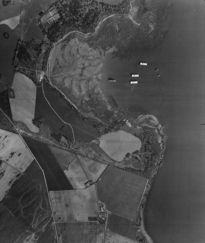 A black and white archive aerial image showing part of a rural coastline. A number of concrete structures can be seen anchored or floating in a sandy bay. 