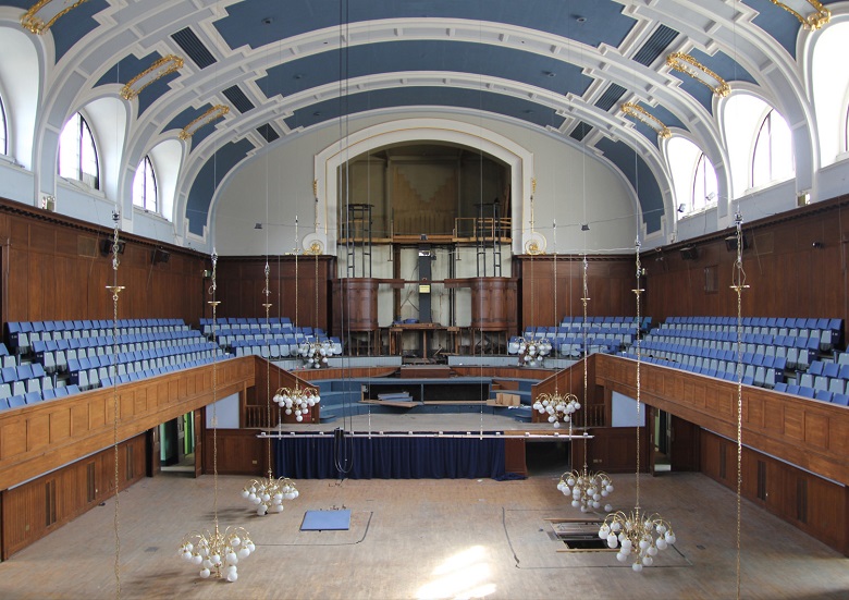 The interior of Perth City Hall prior to recent renovations. There is a stage at one end of the hall, with an empty organ recess behind it. Banks of seats are raised above the main floor on either side. The ornately decorated ceiling is high and curved.