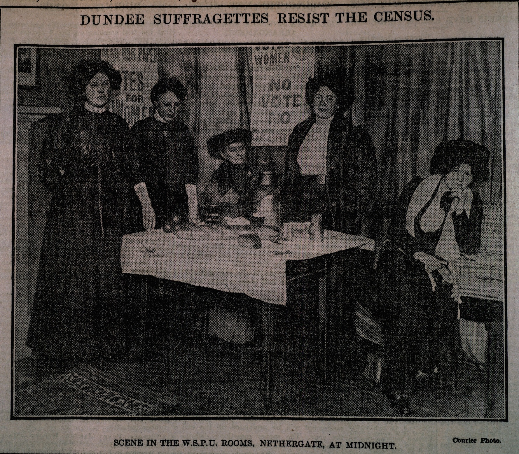 Black and white photo from a newspaper showing suffragettes around a table looking at a camera.