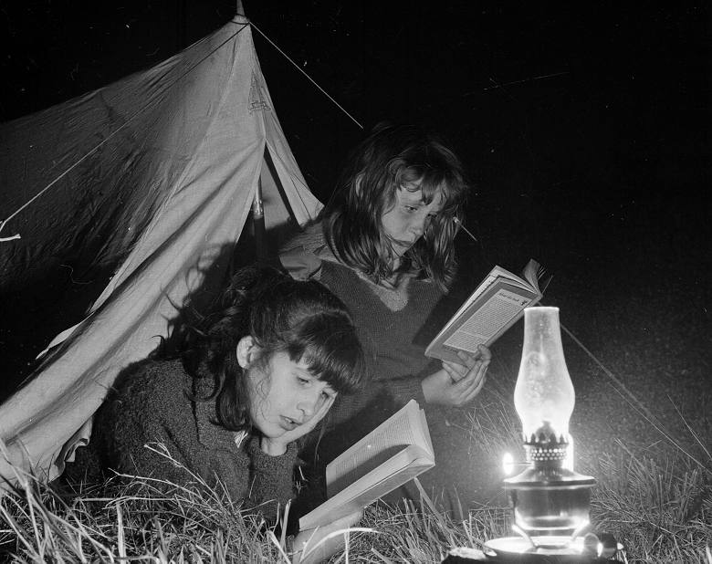 Two girls reading their books by lamplight outside a tent. 