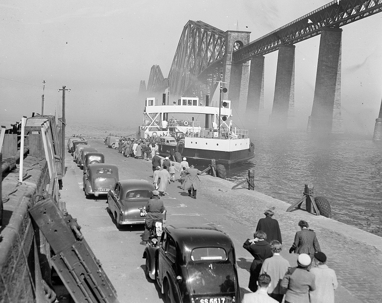 Cars forming an orderly queue on a jetty in the shadow of the Forth Bridge. A car ferry is arriving from the other side of a mist-covered estuary. 