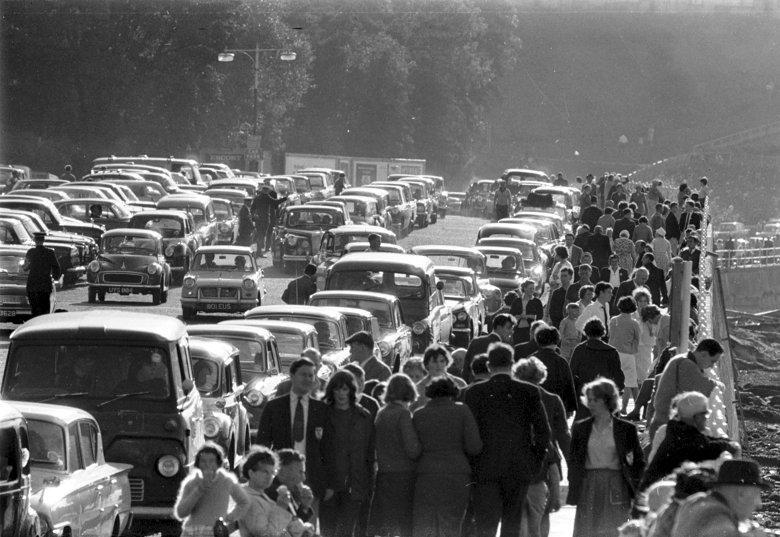 A road full of parked cars and crowds gathering at the side of the River Forth.