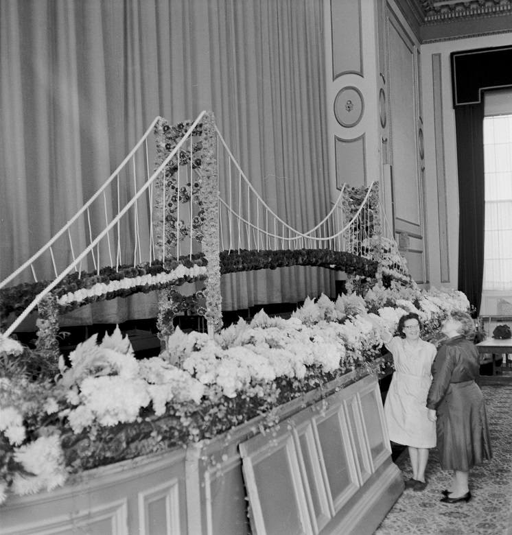 An archive photo of two ladies standing beside a floral model of a suspension bridge on display at a theatre or community hall.