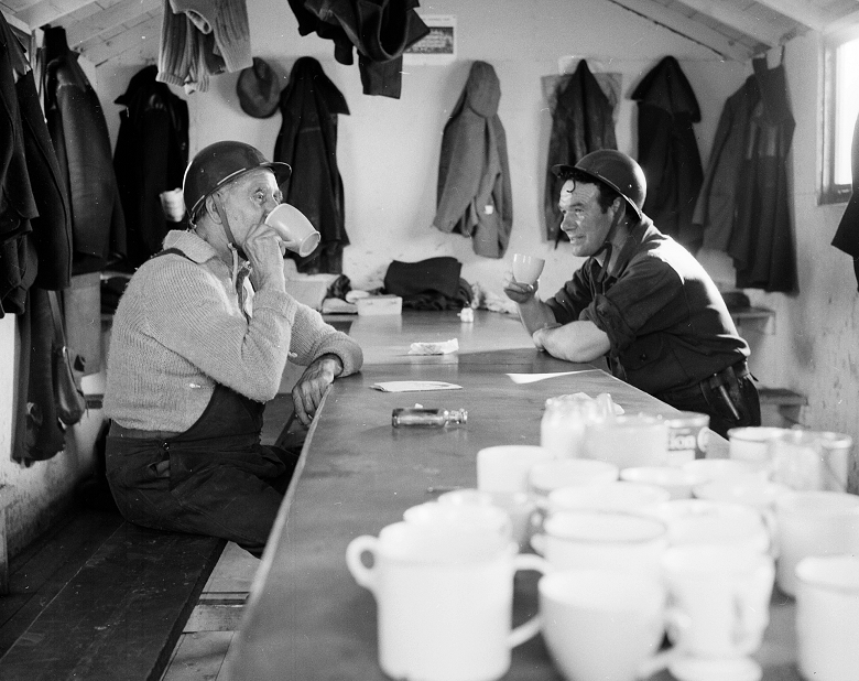 A black and white archive photo of two workmen enjoying a cup of tea at a trestle table in their breakroom. They haven't bothered taking off their hard hats. Various coats and pieces of equipment hang from pegs on the walls. 