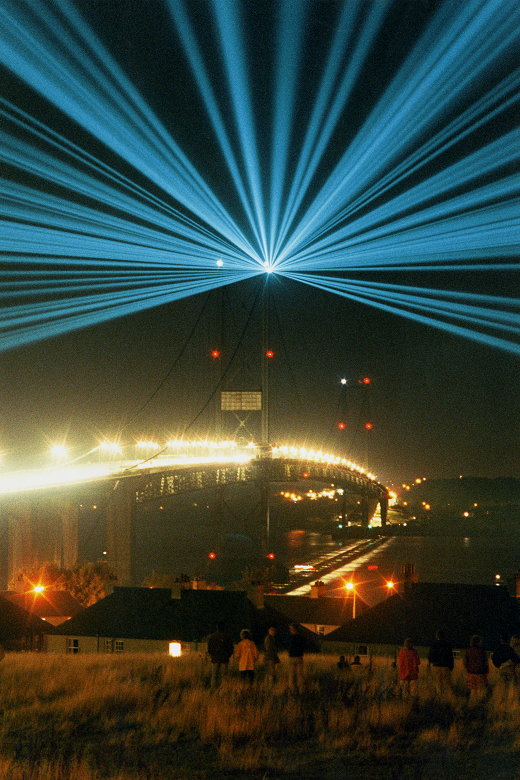 Dazzling glue lasers emitting from the towers of the the Forth Road Bridge as part of a light display. In the foreground spectators look on from a hillside behind houses.