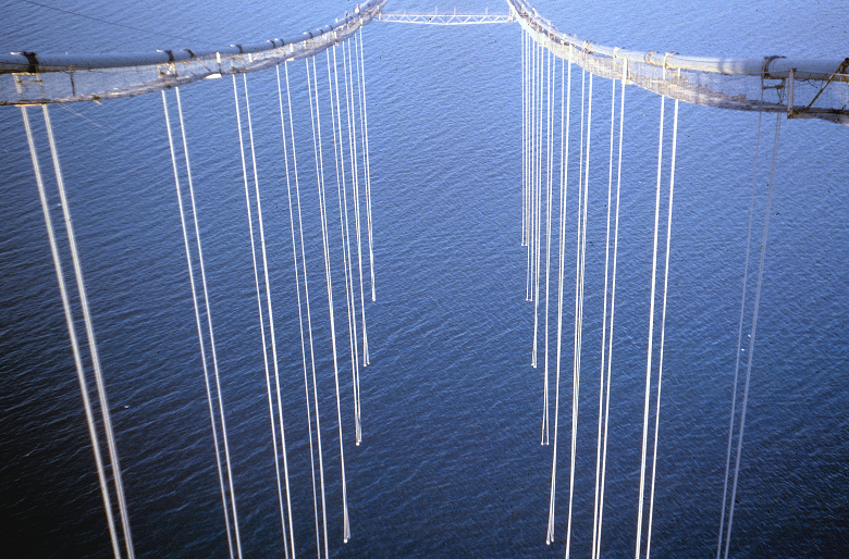 A striking and somewhat unsettling image of a partly constructed suspension bridge. Ropes are hanging from large cables ready to support a roadway. However the road has not yet been built so there is an empty space and a vast expanse of water below.