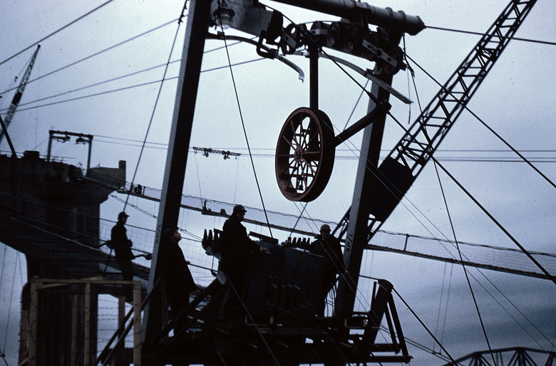 Workers and equipment at the Forth Road Bridge construction site silhouetted against the sky. In the centre of the image is a large wheel, part of a pulley system used to carry wire across the river.