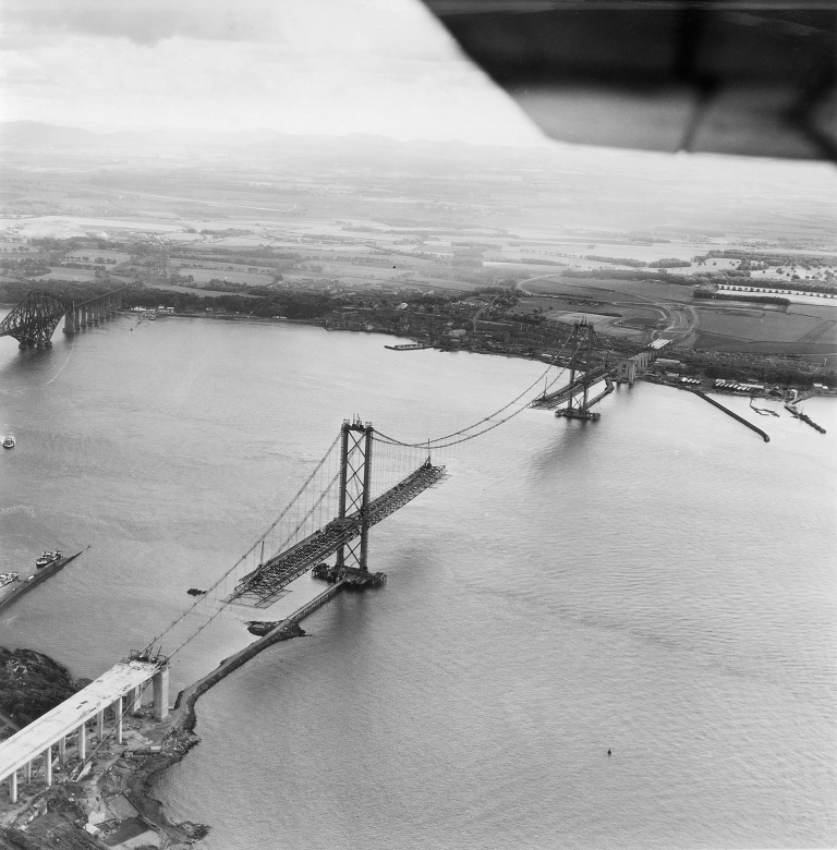 An aerial photo showing a huge suspension bridge under construction. Carriageways are being built outwards from two tall towers and the sections will eventually meet to form a complete crossing. 