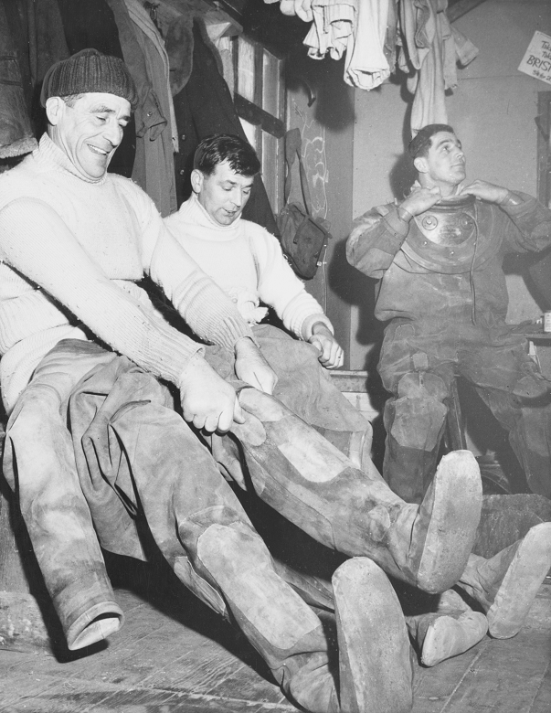 An archive photo of three men pulling on diving suits. They are already wearing several layers of wool jumpers and hats. 