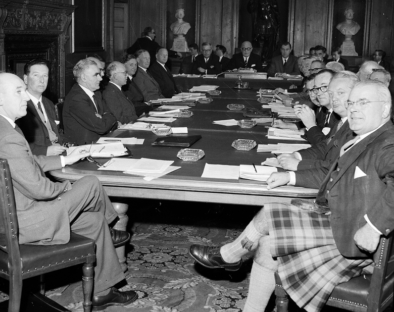 A group of board members gathered around a long table in a grand meeting room. They are dressed smartly in suits and ties and, in one case, a kilt. Every person in the photo is a white man of middle age or older, leaving us in no doubt it was taken in the 1950s.