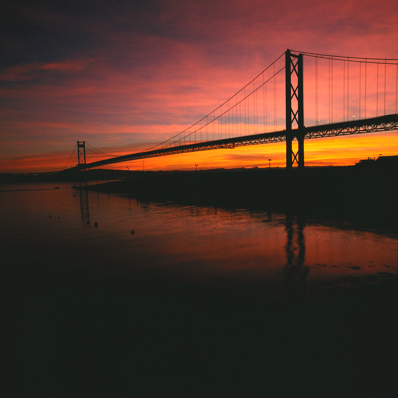 A suspension bridge dramatically silhouetted against a red and pink sky at sunset. In the foreground the waters of a large river a calm and tranquil. 