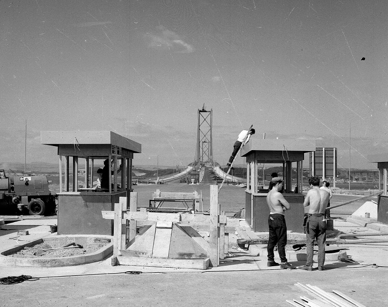 A black and white archive image from the construction of the Forth Road Bridge. On a hot day, two shirtless builders hold a conversation while a third is up a ladder working on a tollbooth. 