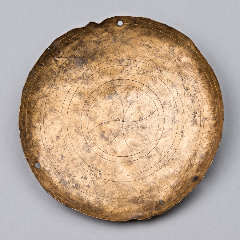 A hammered bronze dish. It's very shallow, almost flat. It is engraved with concentric circles and in the middle is a simple geometric design that looks a bit like a four petalled flower. There are three holes pierced at regular intervals around the edge where it would have hooked on to the arm of the scale.